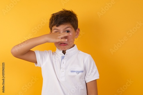 Принти на полотні Displeased little cute boy kid wearing white t-shirt against yellow wall plugs nose as smells something stink and unpleasant, feels aversion, hates disgusting scent
