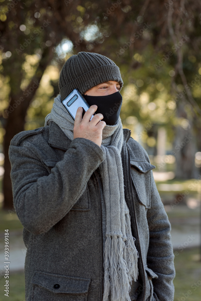 A Adult male wearing a facemask with winter clothes and talking on the phone in a park