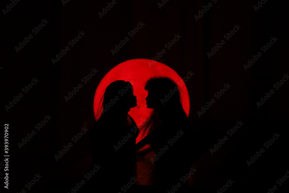 A silhouette of two female dolls standing very close to each other in front of a circular red light source in the dark	