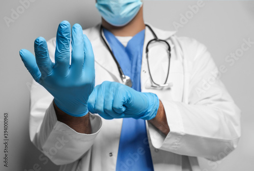 Doctor in protective mask putting on medical gloves against light grey background, closeup