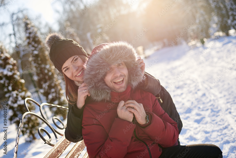 Obraz premium Young couple in love, enjoying winter sunny weather outside in snowy park forest. Brunette woman in black jacket hugging man in red. Christmas and new year holidays joy. Romantic date, relationship.