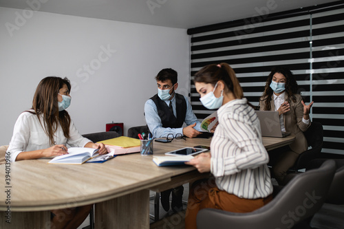 Group of four business people working together in their office on important project. They are wearing face protective masks, talking and analyzing their business plan together. Covid-19 concept.
