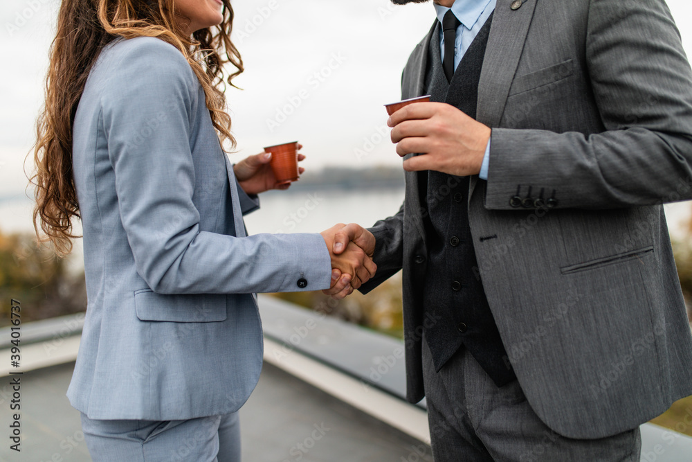 Male and female business colleagues standing on the roof of modern building during work timeout. They are drinking coffee, smiling and talking. Beautiful river landscape in the background.