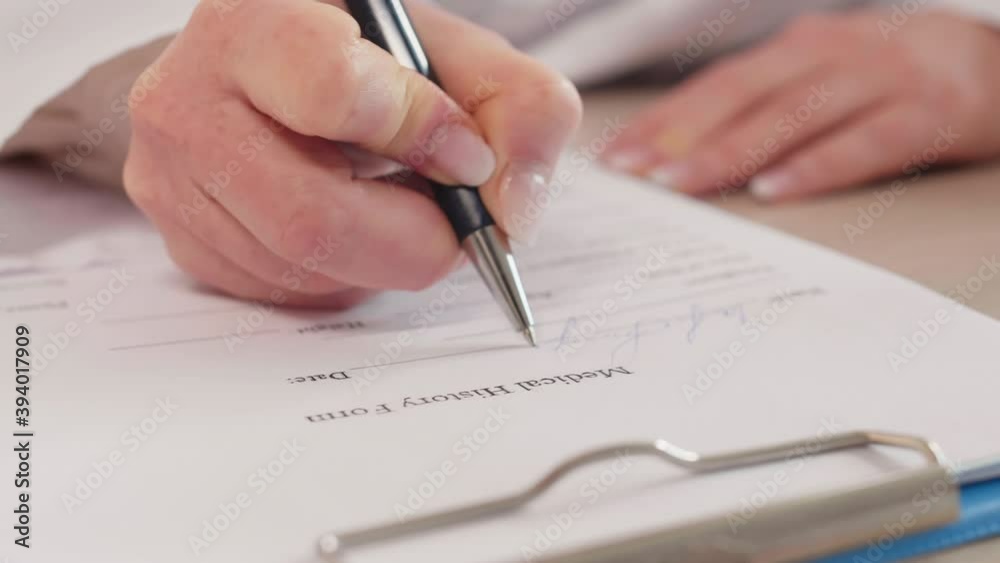 Close-up shot of female hands of unrecognizable doctor writing with pen on paper, filling in medical history form on clipboard Close-up shot of female hands of unrecognizable doctor writing with pen on paper, filling in medical history form on clipboard