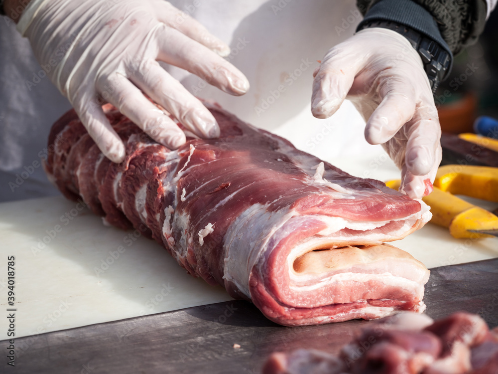 Hands of a professional butcher cutting a piece of raw pork meat with a long knife, on a white plank, at a rural butchery of Serbia. Pork meat is one of the biggest productions in Serbia