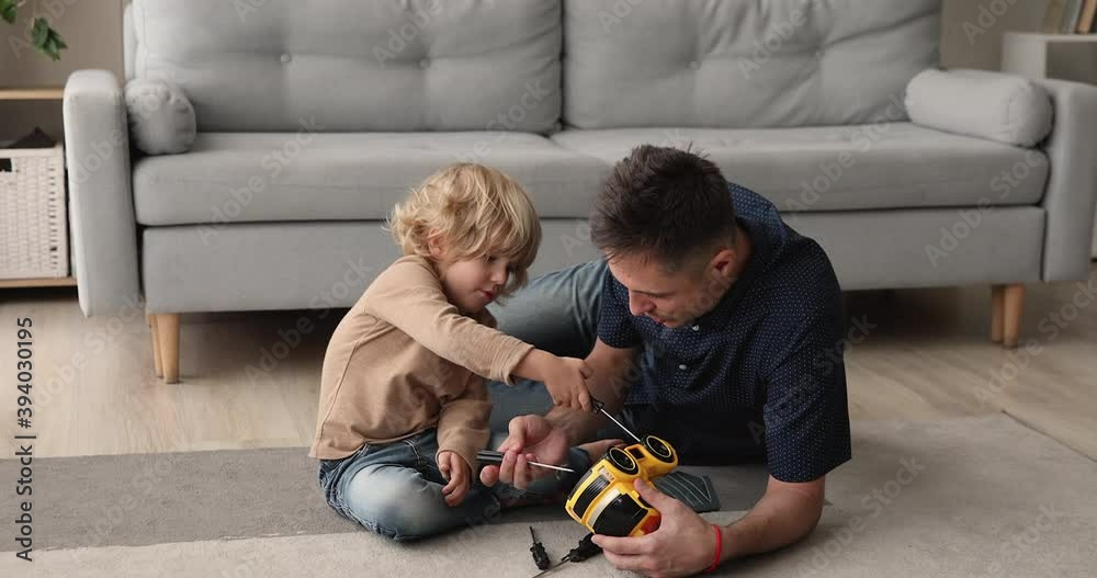 In warm living room young father his little son repairing toy car play together auto mechanic, holding screwdriver fixing broken truck with dad. Skills transmission, share knowledge, boyhood concept