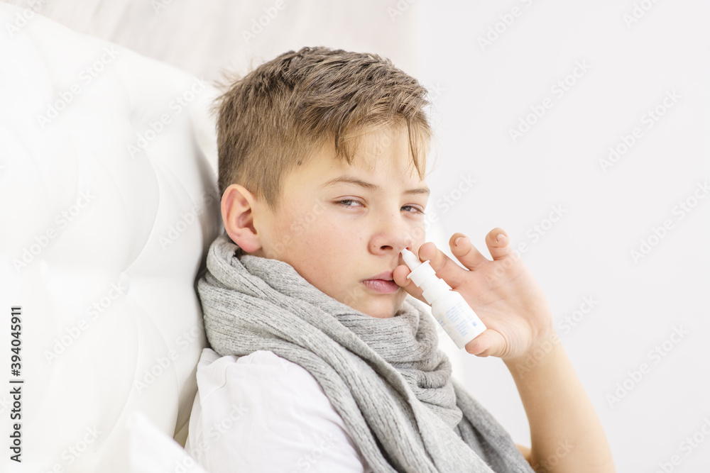 Sick teen boy spray his nose with medicine while sitting on bed at home. Cold season. Sick teen boy spray his nose with medicine while sitting on bed at home. Cold season.