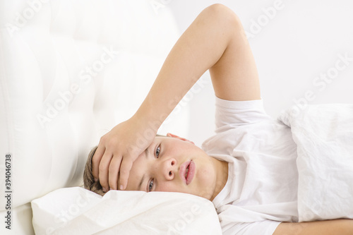 A sick little boy measures the temperature while lying on the bed at home. An unhealthy child holds his hand on his head in bead. Cold season.