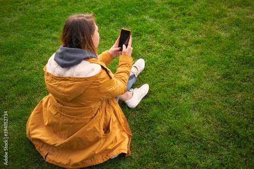 Beautiful pensive woman sits in the park on the grass and takes pictures on a smartphone