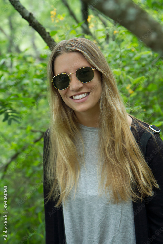 Enjoyment. Portrait of blond young caucasian woman wearing sunglasses and smiling, in the forest. 