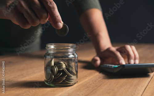 A man put coins in a glass jar and took notes to save money on a wooden table.