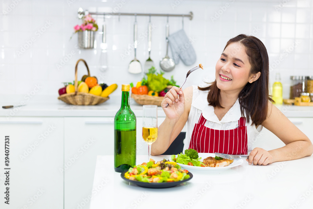 Asian woman eating steak and salad in the kitchen with fruit wine.