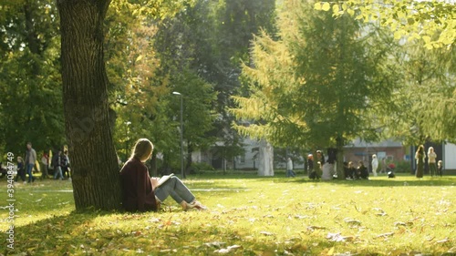 Young unrecognizable woman reading a book in the park. Early autumn, yellow leaves falling on the grass. Sunny and windy day. Steadicam shot