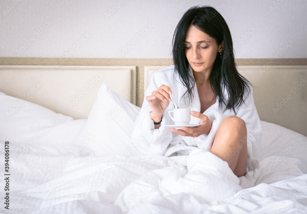 Attractive caucasian brunette woman sits in cosy bed and stirs her morning coffee in espresso cup Attractive caucasian brunette woman sits in cosy bed and stirs her morning coffee in espresso cup