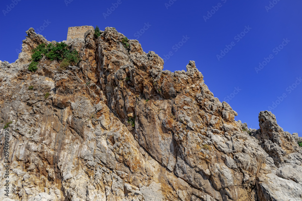 The peak of the rock with the wall of the Alanya Castle at the top (Turkey). Rough stone clump against the blue sky, close-up The peak of the rock with the wall of the Alanya Castle at the top (Turkey). Rough stone clump against the blue sky, close-up