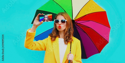 Portrait of attractive young woman taking selfie picture by phone holding colorful umbrella over blue background