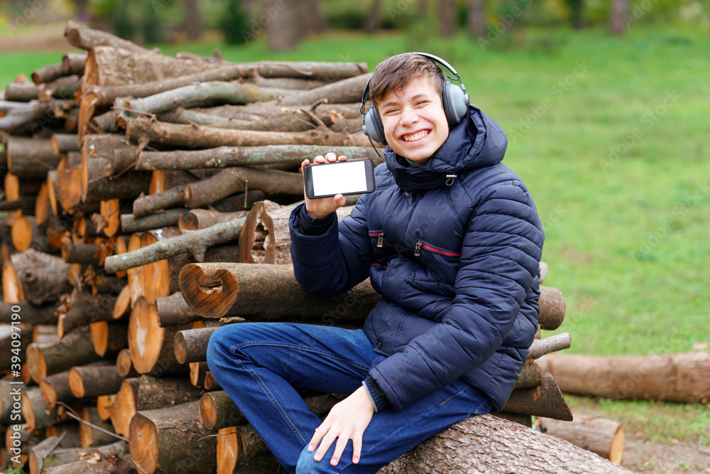 Fototapeta premium teenager listening to music by headphones, relaxing in autumn city park, shows the phone screen and sitting on a log