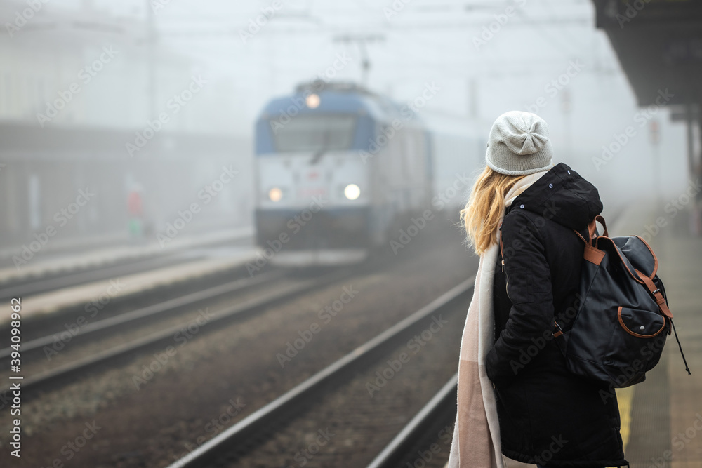 Traveler standing on railroad station platform and looking at arriving train from fog. Woman travel by train in winter