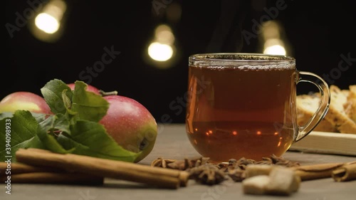Close-up hot tea in glass mug on black background. Steam rises above the cup. Fresh apples with leaves and spices on the table. Slide or move camera. Dolly shot