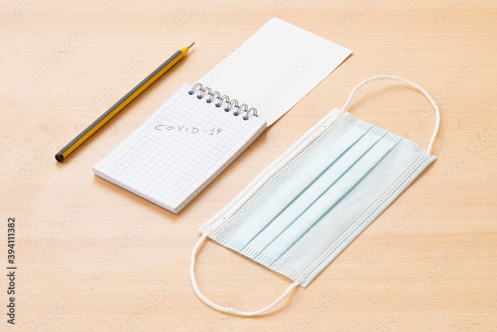 Flat lay, a face mask, a spiral notepad with Covid-19 written on it and a pen are arranged on a wooden table. To-do list concept during the Covid-19 outbreak. Flat lay, a face mask, a spiral notepad with Covid-19 written on it and a pen are arranged on a wooden table. To-do list concept during the Covid-19 outbreak.