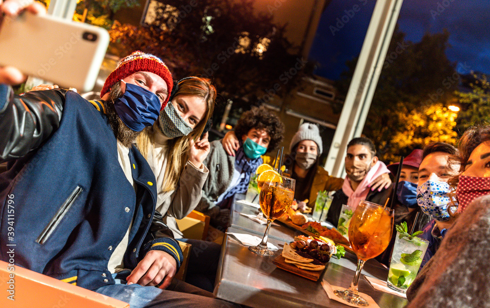 Group of happy friends taking a selfie at cocktail bar - Young people covered by face masks having fun at restaurant - New normal lifestyle concept.