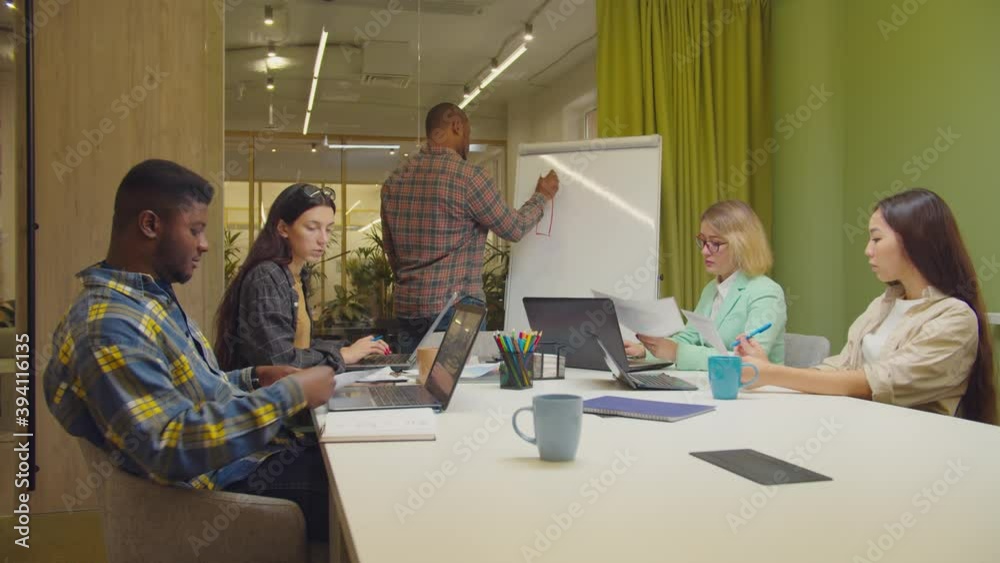 Confident african american male manager drawing financial graph on flipchart, pointing and setting business goals for diverse multiracial business team while working together on project at office.