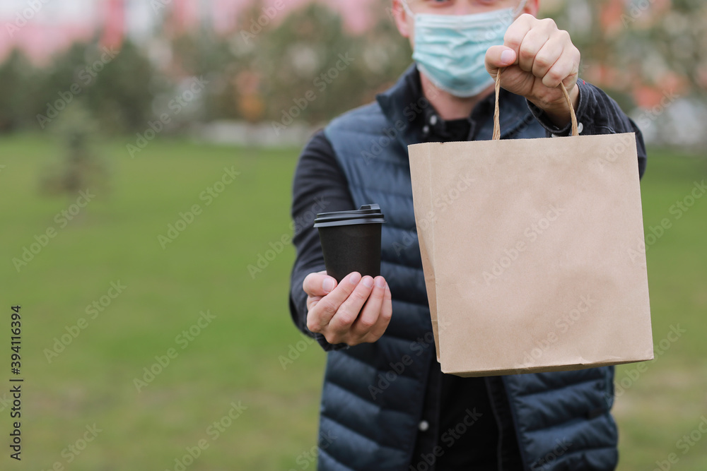 Delivery man in red cap, face medical mask hold take away paper bag and drink in disposable cup outdoors in city. Service coronavirus. Online shopping Delivery man in red cap, face medical mask hold take away paper bag and drink in disposable cup outdoors in city. Service coronavirus. Online shopping