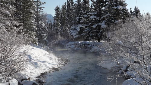 Beautiful tranquil creek in the forest on winter sunny day morning. Mist floating on river surface. Clear blue sky, snow covered pine trees on river bank. Beautiful natural landscape. photo