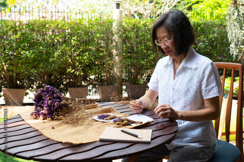 senior asian retired woman make postcard from dry flowers as a hobby. She sits in garden at home