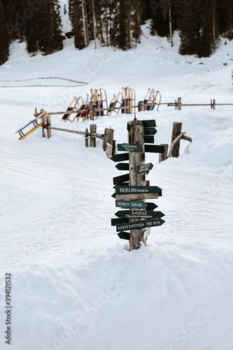 Directional road signs tower standing in the snow in front of sleds parking in the Alps in Italy