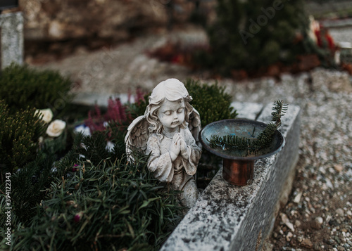 Serene cherub angel statue made with concrete praying on a grave decorated with flowers and plants