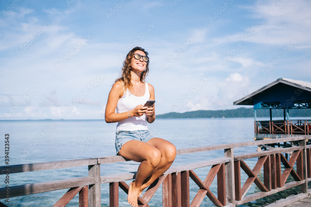 Happy hipster tourist in sunglasses resting at nature environment with seashore on background, cheerful woman with cellphone enjoying summer vacations for international visiting and solo travelling