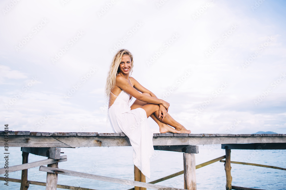 Portrait of cheerful woman in white sundress resting at wooden pier at seashore of Maldive island, carefree Caucasian female tourist visiting resort paradise on Seychelles enjoying recreation