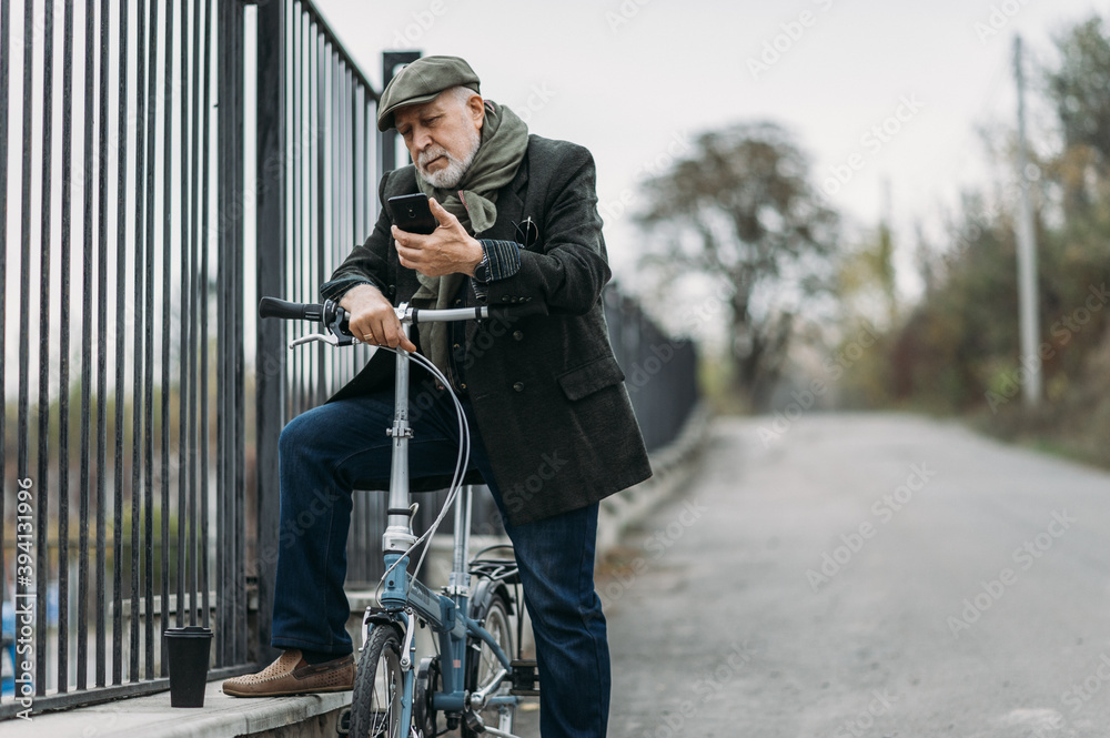 Senior gray-haired business man using smartphone app while sitting on bike with a cup of coffee - Hipster businesswoman driving to work - road, morning break and tech concept - Focusing on face