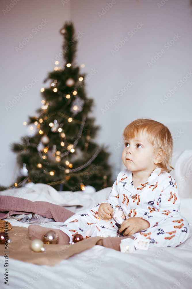 the child's first Christmas. a pensive little boy is lying in his holiday pyjamas on a bed against the background of a brightly decorated fir tree with a Golden light garland.