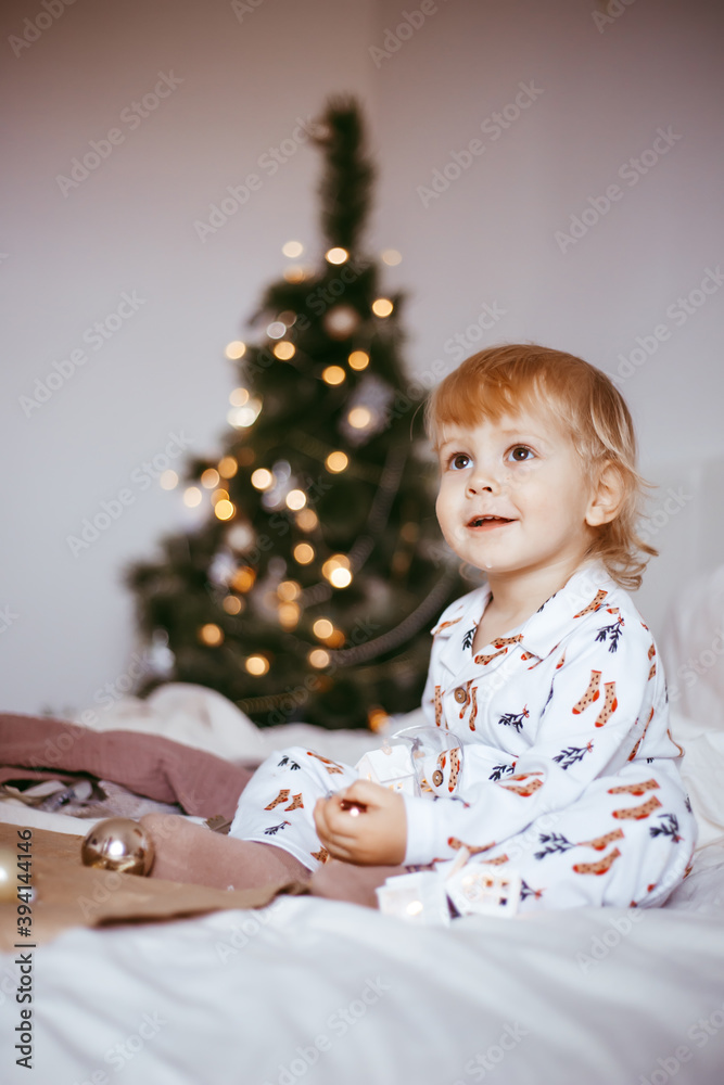 the child's first Christmas. a pensive little boy is lying in his holiday pyjamas on a bed against the background of a brightly decorated fir tree with a Golden light garland. the child's first Christmas. a pensive little boy is lying in his holiday pyjamas on a bed against the background of a brightly decorated fir tree with a Golden light garland.