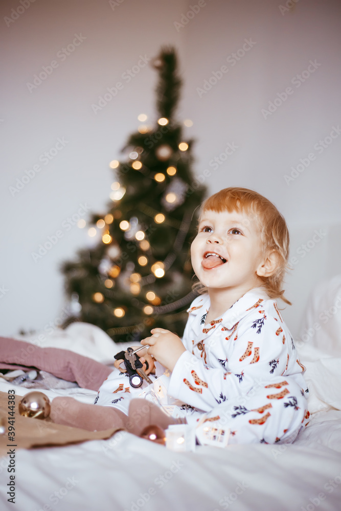 the child's first Christmas. a pensive little boy is lying in his holiday pyjamas on a bed against the background of a brightly decorated fir tree with a Golden light garland. the child's first Christmas. a pensive little boy is lying in his holiday pyjamas on a bed against the background of a brightly decorated fir tree with a Golden light garland.