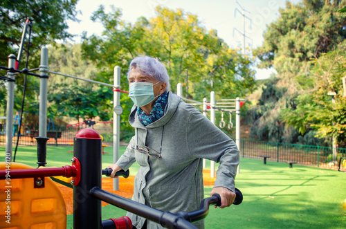 Fototapeta Naklejka Na Ścianę i Meble -  A senior adult woman in a face mask starts exercising at an outdoor fitness station near her residence. She knows that physical activity boosts the immune function elderly people.