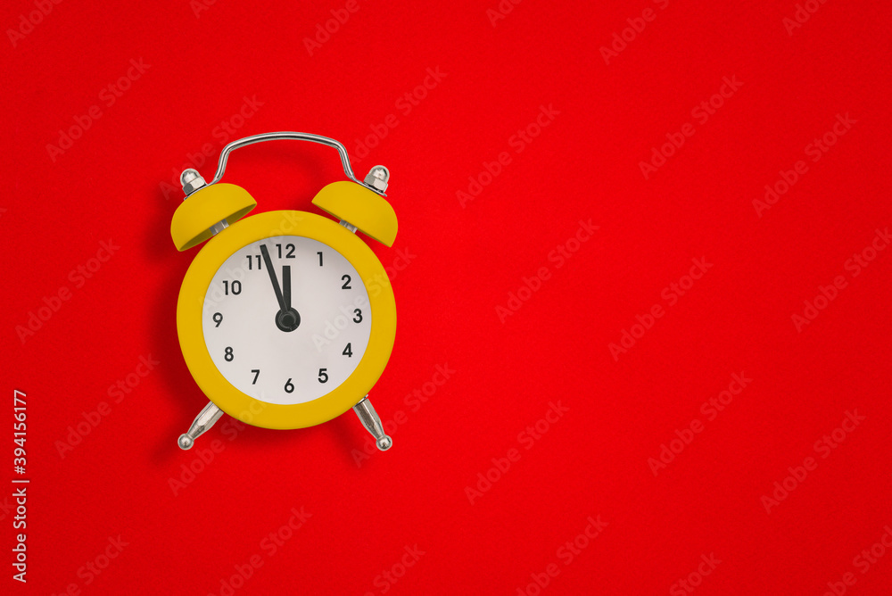 Yellow alarm clock on red paper with copy space. The hands of the clock show twelve o'clock Yellow alarm clock on red paper with copy space. The hands of the clock show twelve o'clock
