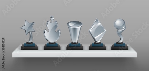 Realistic bookshelf with trophies. 3D transparent glass awards on black stands. Blank winner prizes, creative and scientific competition cups. Collection of sport champion reward. Vector victory set