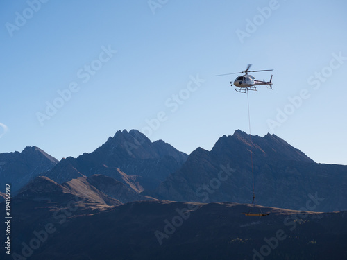 A mountain scene with a helicopter flying and transporting materials that hang from a cable attached to the helicopter.