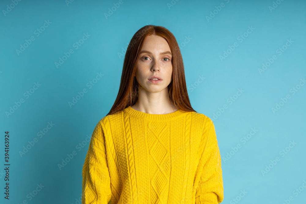 Fototapeta premium redhead beautiful caucasian young woman with healthy freckled clean skin, in yellow sweater looking at camera with calm and serious expression om blue background. Closeup studio shot