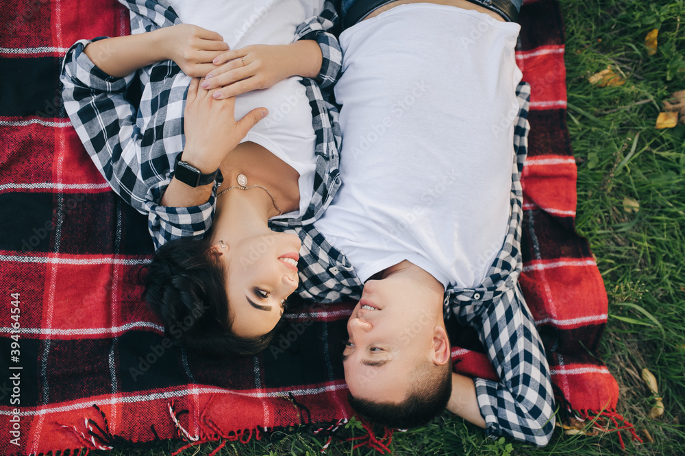 A beautiful, smiling and loving guy and a girl in a plaid shirt and jeans lie on a red plaid in the fall in yellow leaves. Photo, top view.