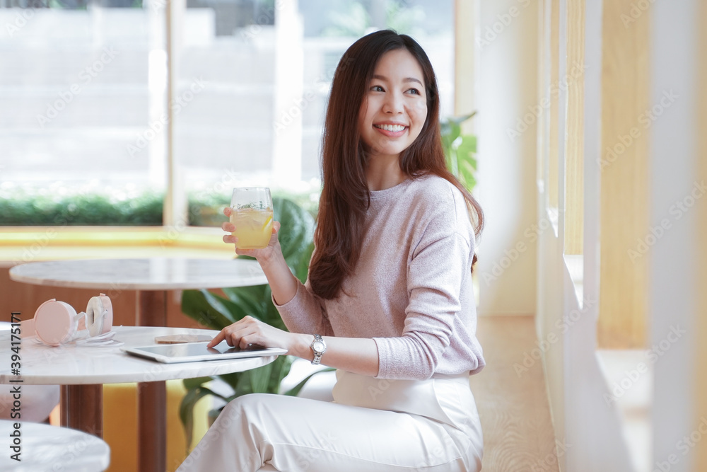 Young Asian business woman in casual clothes smile and sitting in cafe holding a glass of juice and looking out the window with tablet and pink headphones on the table Young Asian business woman in casual clothes smile and sitting in cafe holding a glass of juice and looking out the window with tablet and pink headphones on the table