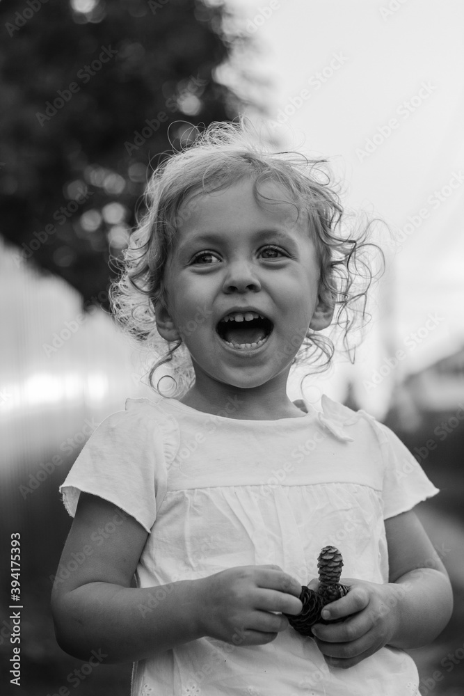 A little blond curly girl is standing near the fence in the countryside in summer with laugh and open mouth black and white, vertical - obrazy, fototapety, plakaty 