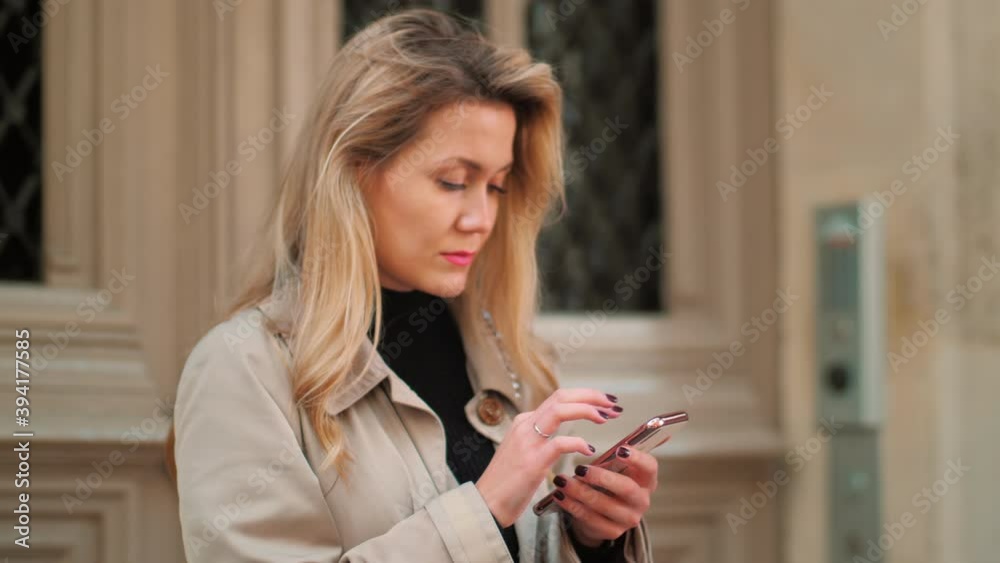 Blonde in a beige trench in Paris uses a smartphone in the background of the building. High quality 4k footage Blonde in a beige trench in Paris uses a smartphone in the background of the building. High quality 4k footage