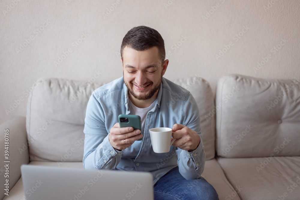Happy man drinking a cup of coffee at home. Shot of a young man using a smartphone and laptop and having coffee on the sofa at home. Work at home. Happy man drinking a cup of coffee at home. Shot of a young man using a smartphone and laptop and having coffee on the sofa at home. Work at home.