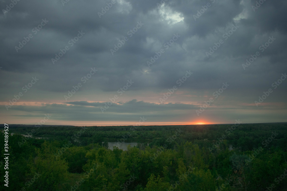 The sun almost went down the horizon, cloudy gray blue sky, forest and river in the distance