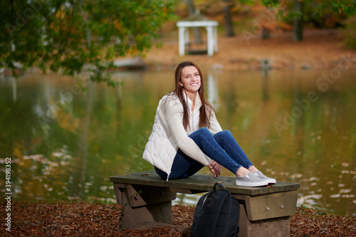 Beautiful young college student sits alone on campus in fall leaves near small pond