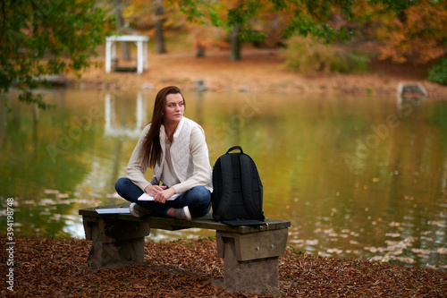 Beautiful young college student sits alone on campus in fall leaves near small pond - writing in notebook